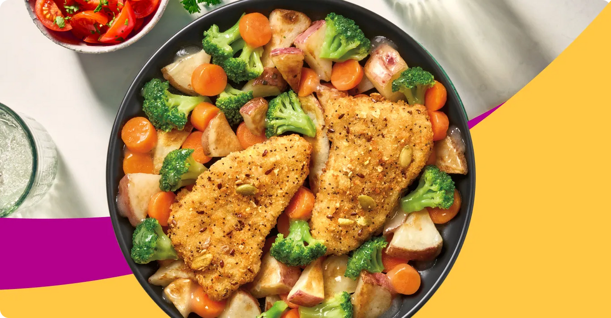 Overhead view of a black skillet filled with two breaded chicken breasts and mixed vegetables including broccoli, carrots, and potatoes, set on a white surface with a colorful yellow and magenta graphic background.