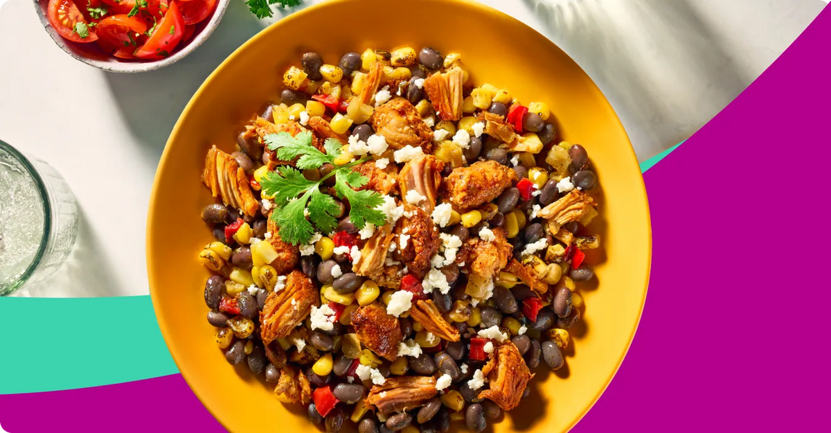 A top-down view of a colorful black bean and corn salad with chicken, feta cheese, and cilantro, served in a bright yellow bowl on a white table with decorative colorful background shapes.