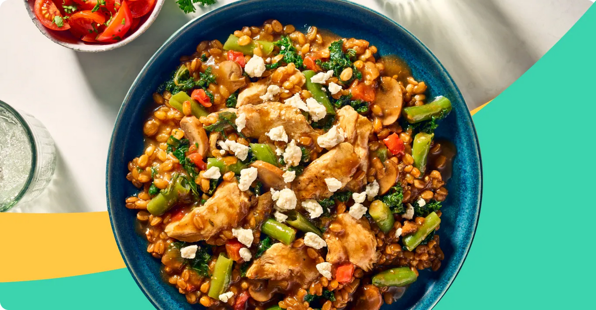 A top-down view of a hearty farro and chicken stew in a dark blue bowl, garnished with crumbled white cheese and fresh green beans, with a small side of sliced tomatoes in the upper left corner.