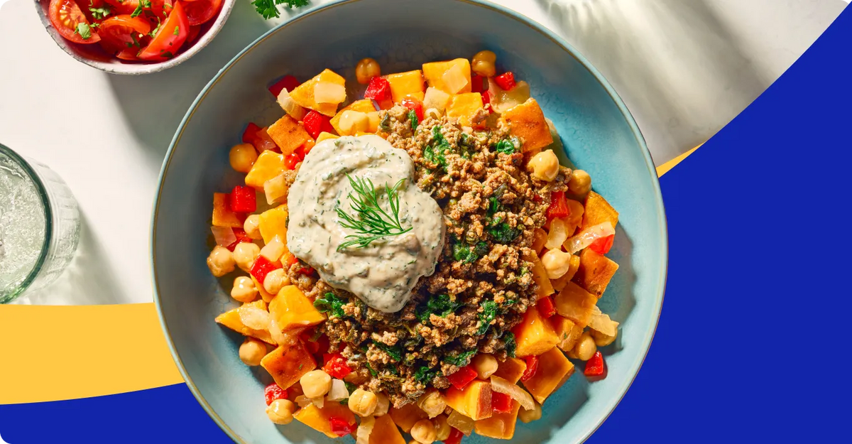 A top-down view of a light blue bowl filled with a colorful chickpea salad, featuring sweet potato cubes, red bell peppers, ground meat, and a dollop of white dressing topped with dill. A small side dish of sliced tomatoes is visible in the upper left corner.